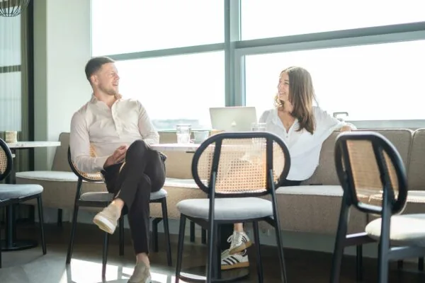 Twee mensen zitten lachend te praten in een moderne lounge op Joop Geesinkweg, met een laptop op tafel.